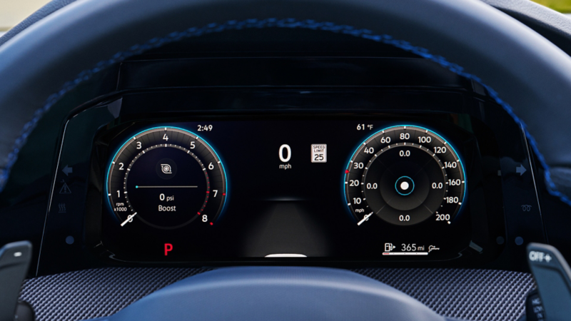 Close-up of the Volkswagen Golf R Digital Cockpit display showing performance gauges, including tachometer, boost pressure, speedometer, and gear position, framed by blue-stitched steering wheel and carbon-style dashboard trim.