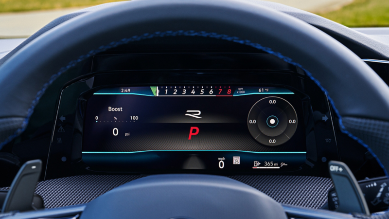 Close-up view of the fully digital instrument cluster inside the Golf R. The screen displays performance metrics including turbo boost pressure (0 psi), RPMs in a horizontal tachometer layout (from 0 to 8,000 rpm), gear indicator ("P"), and fuel range (365 miles). The signature "R" logo is centered, framed by carbon-fiber trim and contrast-stitched leather on the steering wheel.
