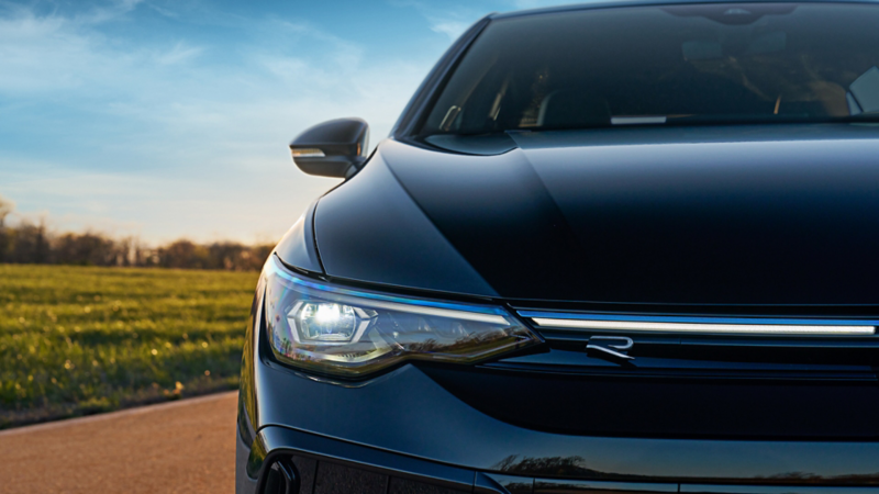 Close-up of the front end of a Volkswagen Golf R in Mythos Black Metallic, highlighting the illuminated right side headlight, R badge on the grille, and sharp body lines at sunset.