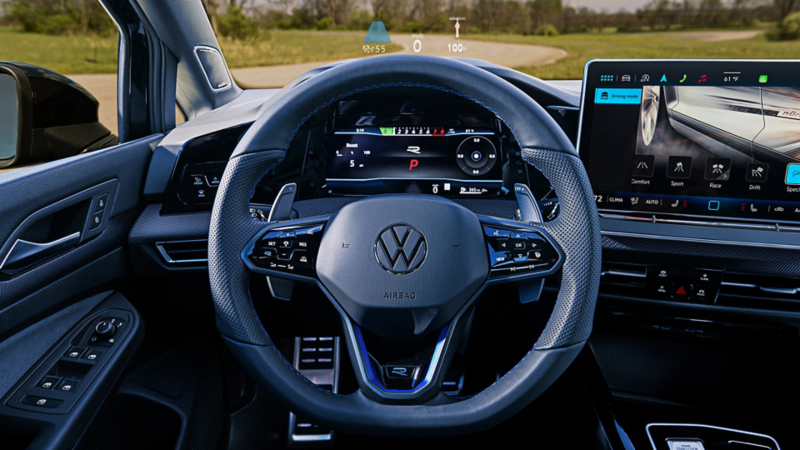 Interior view of the Mythos Black Metallic Golf R showing the steering wheel, fully digital gauge cluster, touchscreen display with drive mode, and head-up display.