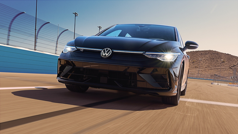 Front view of a Volkswagen Golf R in Mythos Black Metallic driving on a racetrack, captured low and close-up from the front during a motion shot.