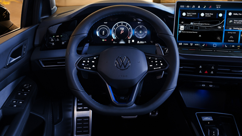 Interior view of the Volkswagen Golf R driver cockpit showing the R-branded steering wheel, digital instrument cluster, large display screen and metallic sport pedals.