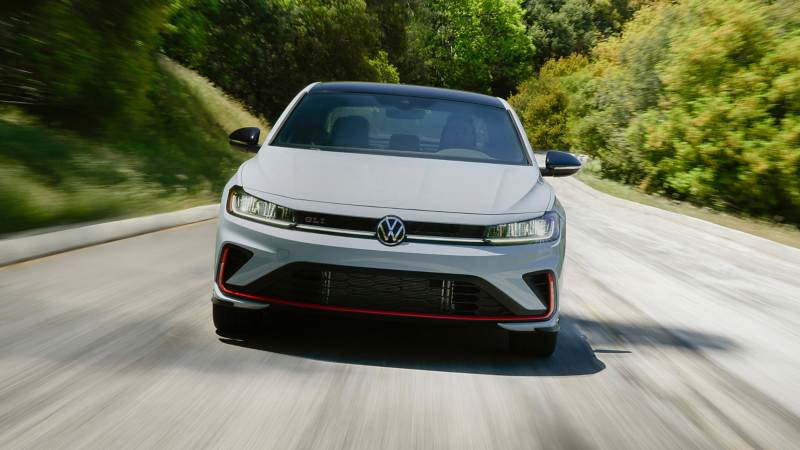 Head-on view of a Jetta GLI shown in Monument Gray driving on an open road with trees in the background