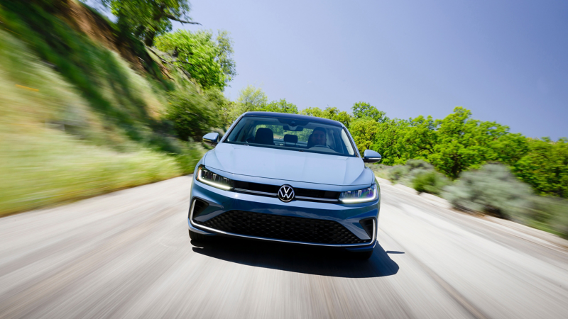 Head-on view of a Jetta shown in Monterey Blue Pearl driving on an open road with trees in the background