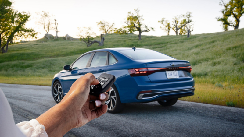 Rear view of a Jetta shown in Monterey Blue Pearl parked with trees in the background and a person holding a VW key fob in the foreground