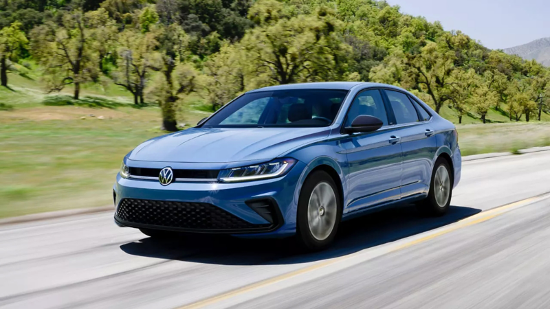 Front ¾ view of a Jetta shown in Monterey Blue Pearl driving on an open road with wooded hills in the background