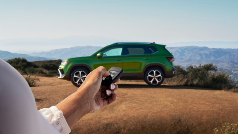 A hand holding a VW key fob with a Taos shown in Bright Moss Green parked in the background.