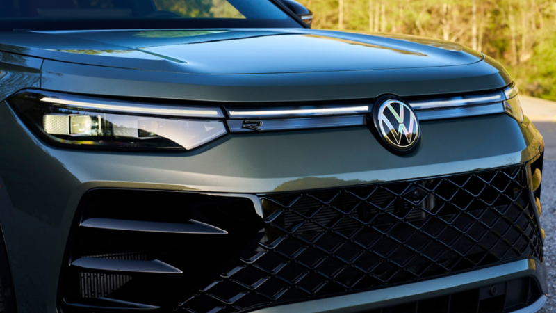 Close up view showing the LED headlights and grille of a Tiguan shown in Avocado Green Metallic with trees in the background.