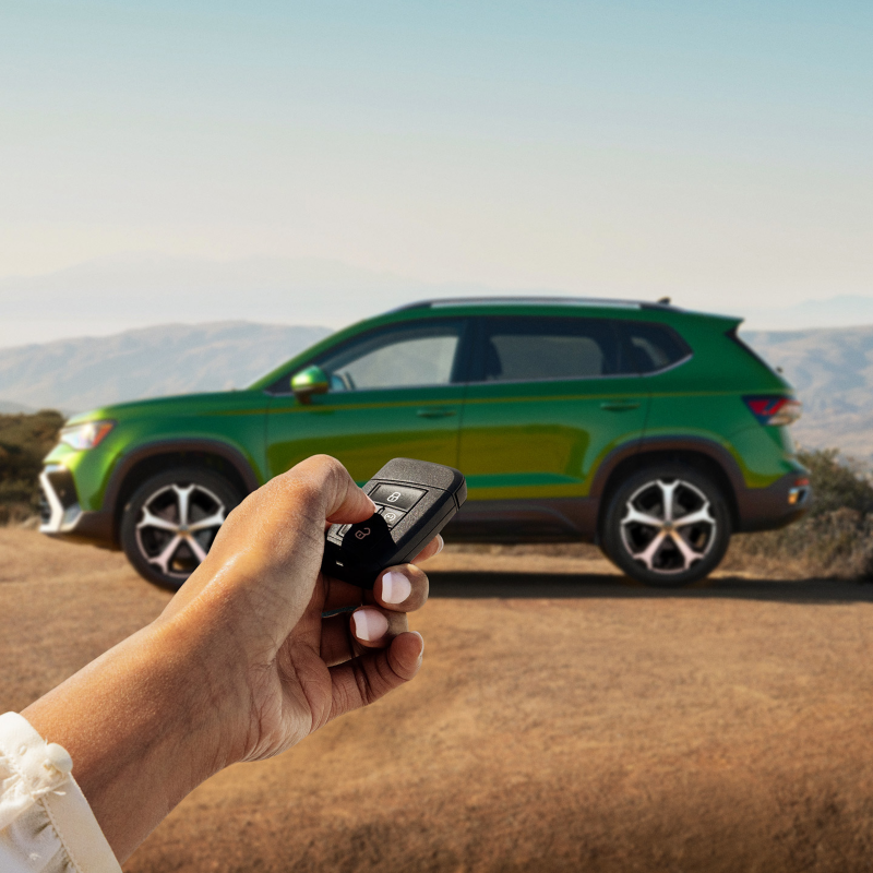 Una mano sostiene un llavero de VW con un Taos en color Bright Moss Green estacionado al fondo.