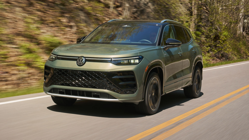 Front 3/4 view of a Tiguan shown in Avocado Green with black roof driving on a mountain road with grassy mountains in the background.