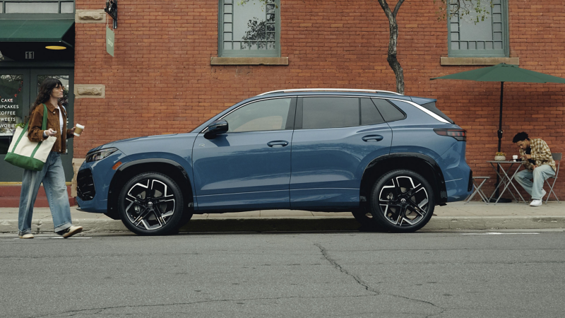 A hip dressed woman carrying coffee walks toward a Tiguan shown in Monterey Blue Pearl parked outside a coffee shop.