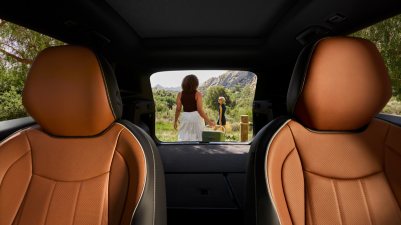 Interior view looking out the open hatch of a parked Tiguan shown in Amber Brown leather with two people holding picnic gear seen outside the vehicle in a wooded park setting.