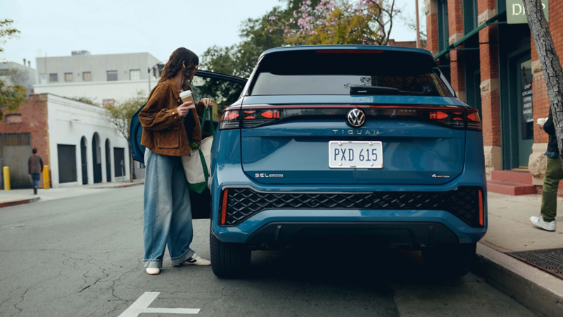 A hip dressed woman carrying coffee opens the driver's door to get in a Tiguan shown in Monterey Blue Pearl parked outside a coffee shop. 