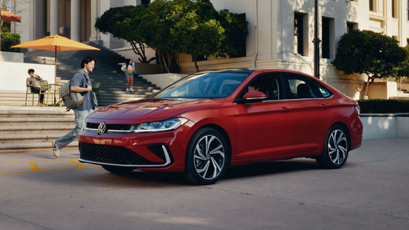 Un Jetta mostrado en Kings Red Metallic está parqueado en un campus universitario mientras un estudiante camina hacia el Jetta.