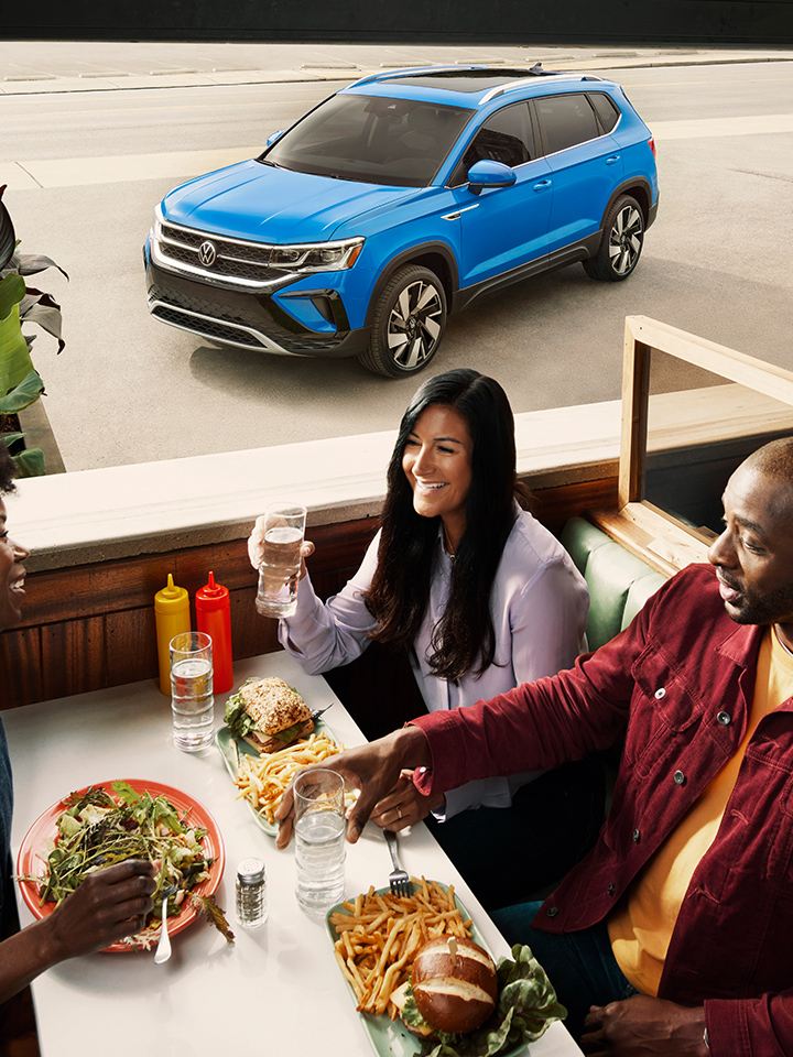 Three people seated in a booth at a diner, as the front driver side of a Taos in Cornflower Blue is seen through the window.