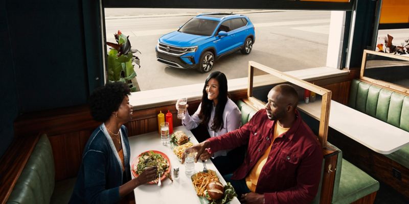 Three people seated in a booth at a diner, as the front driver side of a Taos in Cornflower Blue is seen through the window.