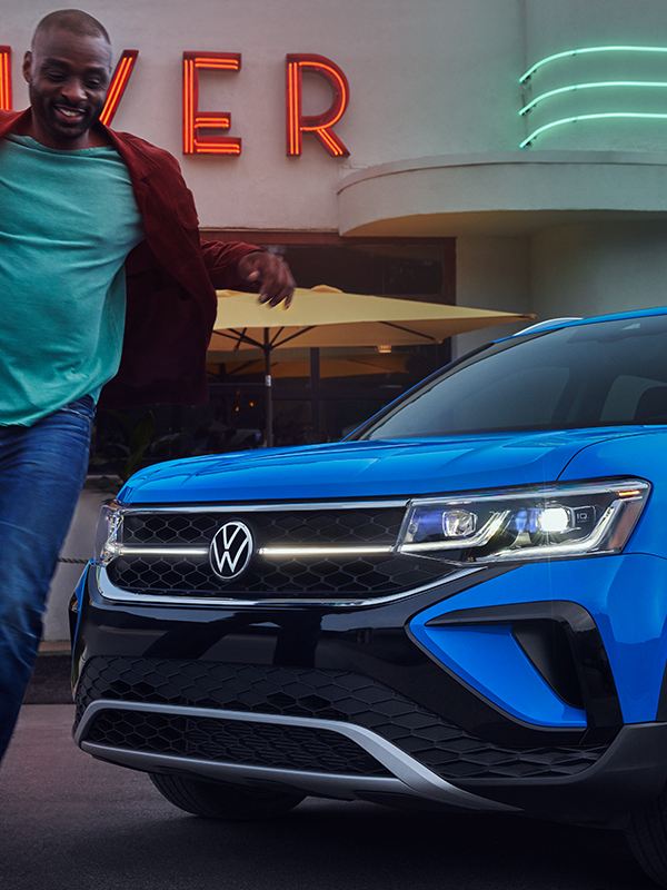 Close-up shot of a man walking in front of a Taos in Cornflower Blue parked in front of a neon-accented restaurant named “Pearl Diver.”
