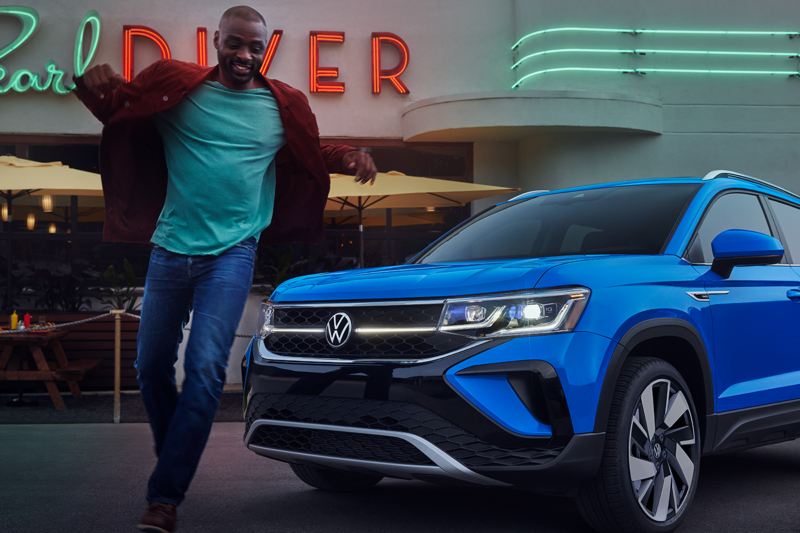 Close-up shot of a man walking in front of a Taos in Cornflower Blue parked in front of a neon-accented restaurant named “Pearl Diver.”