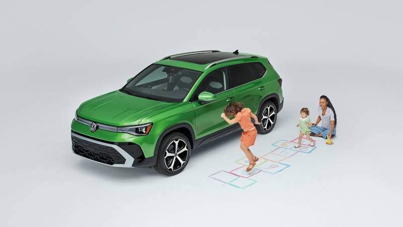 Overhead view of a mom sitting on the ground while children play next to a Taos shown in Bright Moss Green parked on a white sound stage environment.