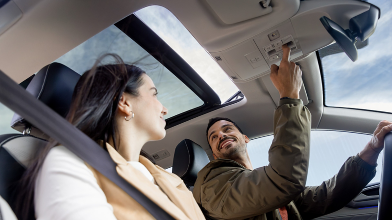 Two smiling people sitting in the front seats of a parked Taos while the driver uses the overhead console controls to open the available panoramic sunroof.