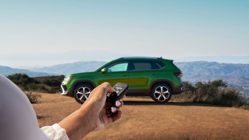 A hand holding a VW key fob with a Taos shown in Bright Moss Green parked in the background.