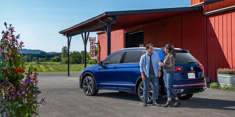 A couple talks at the rear driver side of a Tiguan shown in Atlantic Blue Metallic outside a roadside farm.