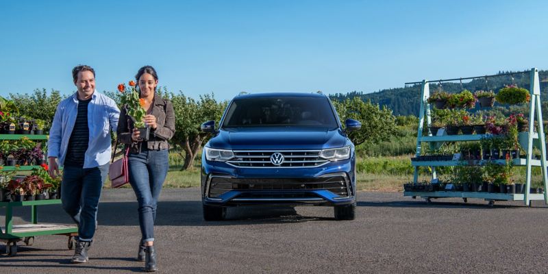 Una pareja sonriente, que lleva una pequeña maceta con flores, compra en una granja al lado de la carretera, cerca de un Tiguan estacionado en color Atlantic Blue Metallic.