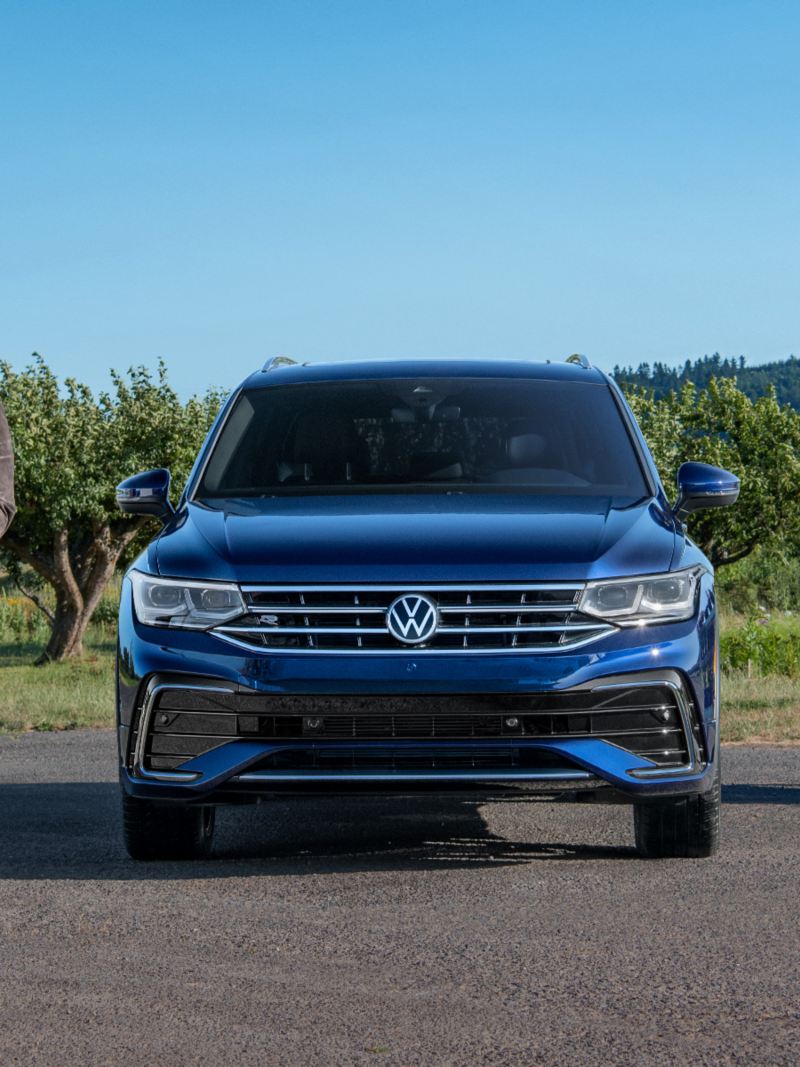 A smiling couple, carrying a small potted flowering plant, shops at a roadside farm near a parked Tiguan shown in Atlantic Blue Metallic.