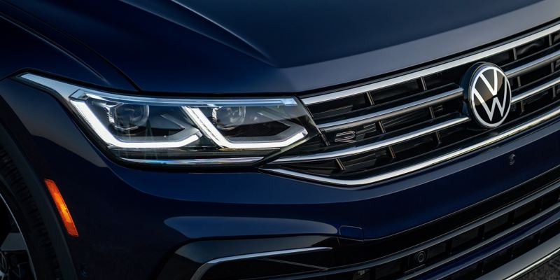 Close up shot of LED headlights and R-Line badge on the front grille of a Tiguan shown in Atlantic Blue Metallic.