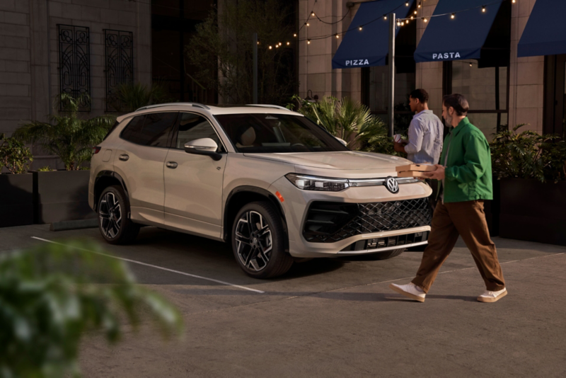 Vista frontal de 3/4 de un Tiguan en color Sandstone estacionado frente a un restaurante, con una persona caminando hacia el vehículo mientras lleva comida.