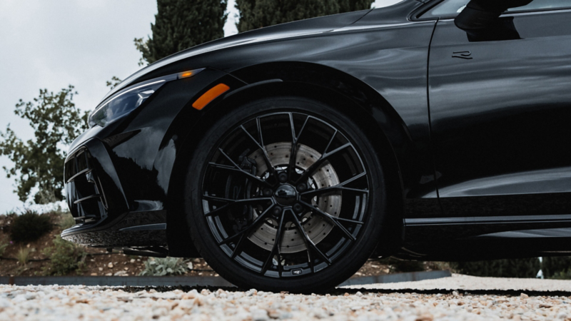 Close up profile view of the front driver side wheel on a Golf R shown in Mythos Black Metallic parked with trees in the background.