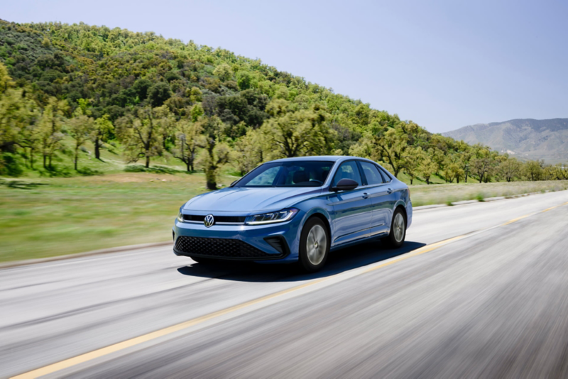 Front ¾ view of a Jetta shown in Monterey Blue Pearl driving on an open road with wooded hills in the background.