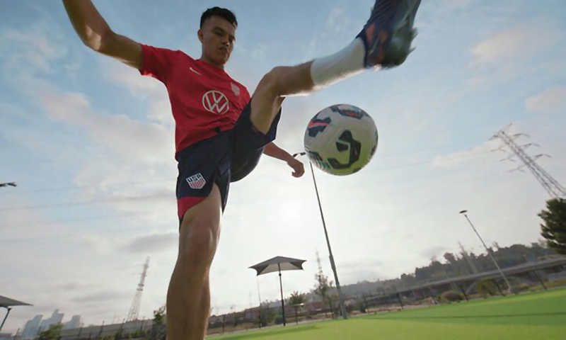 A soccer player on a grass field is mid-jump kicking a soccer ball in the air.