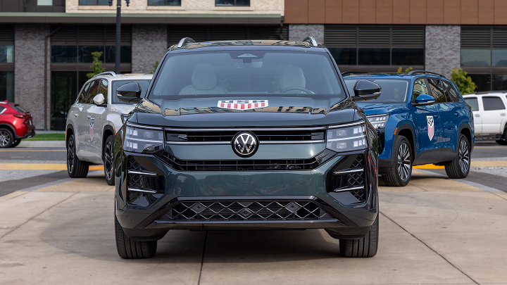 Una vista frontal de un SUV Atlas 2027 en verde Sacramento con el escudo de la Federación de Fútbol de EE. UU. estampado en el capó del vehículo está parqueado delante de otros dos SUV Atlas 2027 visto desde un ángulo frontal, cualquiera de ellos en los colores Silver Mist Metallic y Mountain Lake Blue Metallic. Al fondo hay modernos edificios industriales. 