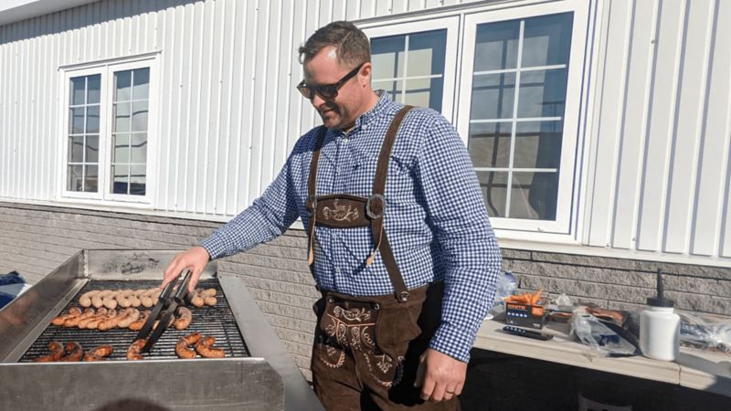 A man wearing lederhosen and sunglasses tends to a barbeque.
