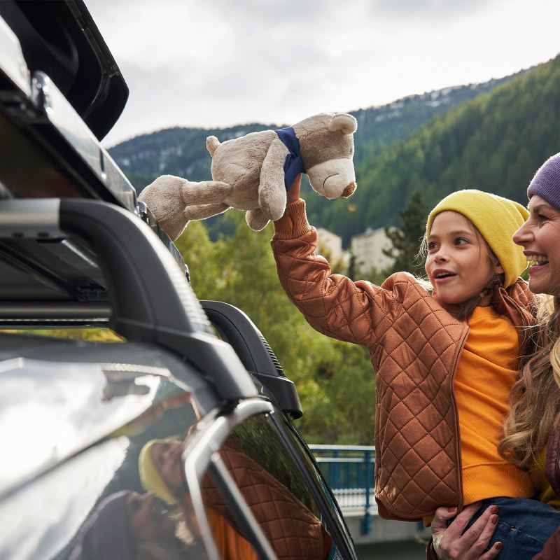 Una bambina, in braccio alla madre, gioca con un orsacchiotto di peluche, vicino alla barre portatutto Originali, montate sul tettuccio di una Volkswagen.