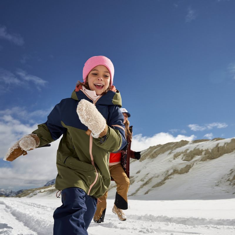 Una bambina, con quanti e cappello di lana, corre su una pista da sci ridendo, inseguita dalla madre