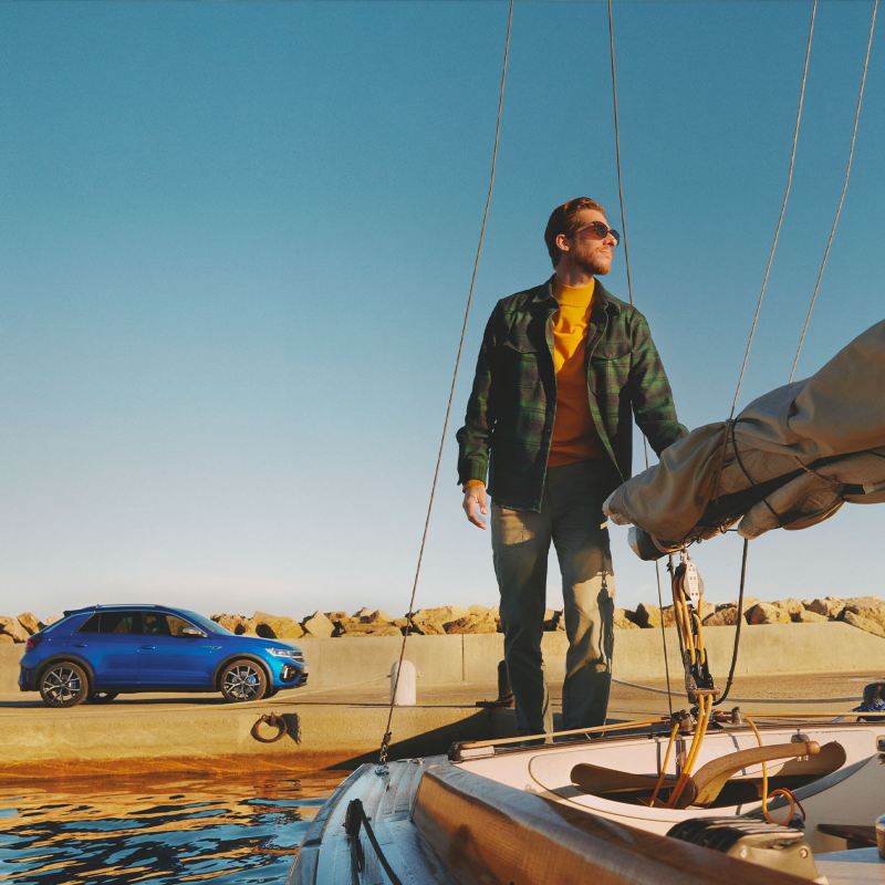 Un uomo si allontana a bordo di una barca a vela, sul fondo è parcheggiata una Volkswagen