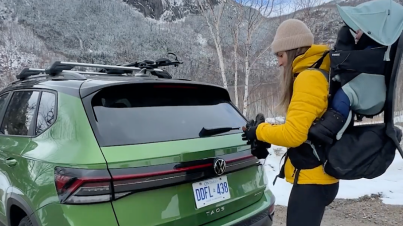 Audrey is standing behind her 2025 Taos with her baby strapped to her back. Snow and leafless trees fill the background.