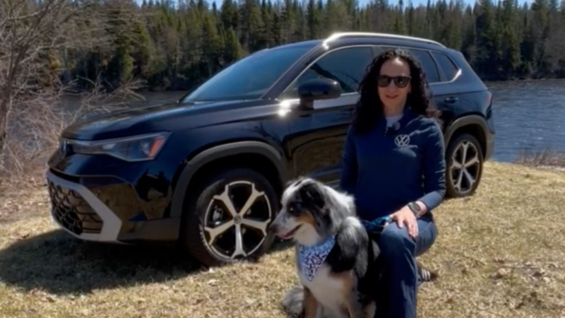 Caroline is kneeling down next to her dog on a field of grass next to a lake. Behind them is a black Taos.