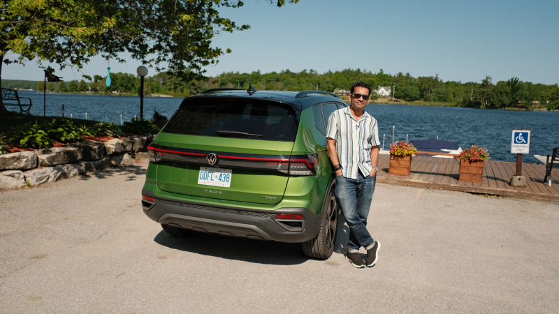 Navin is leaning on a green Taos, posing for the camera on a sunny day. The car is parked next to a boardwalk along a lake. 