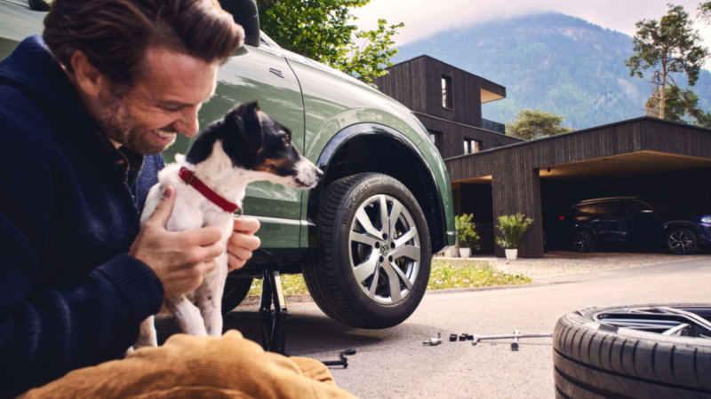 Un hombre sostiene a un perro cerca de un vehículo VW verde, exhibiendo neumáticos de invierno.