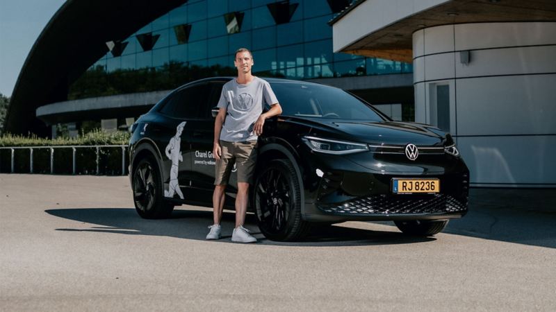 Un homme souriant, vêtu d'un t-shirt et d'un short, se tient à côté d'une Volkswagen ID.5 noire. La voiture est garée devant un bâtiment moderne doté de grandes baies vitrées.