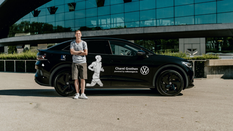 Un homme souriant, portant un t-shirt et un short, se tient les bras croisés à côté d'une Volkswagen ID.5 noire. La voiture est garée devant un bâtiment moderne aux grandes baies vitrées.