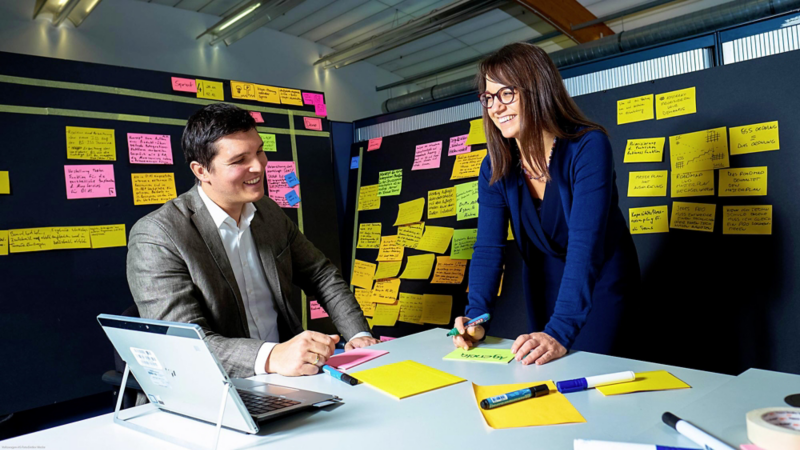Two VW employees chatting happily at a table in front of walls covered with colorful sticky notes