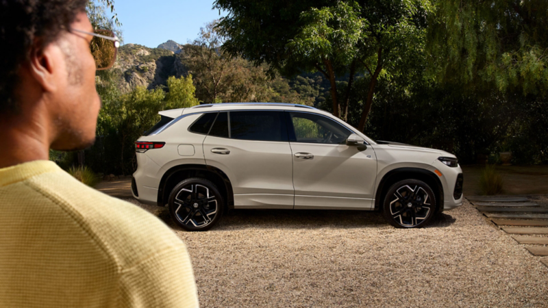 Person looking toward a white SUV parked in a serene outdoor setting with trees and mountains in the background, highlighting a modern design and adventurous lifestyle.