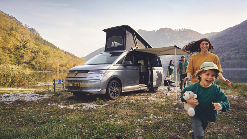 Une famille s'amuse au bord d'un lac à côté d'un van Volkswagen gris avec son toit relevable déployé.