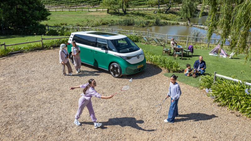 Une famille profite d'une journée en plein air près d'un van électrique Volkswagen ID. Buzz vert et blanc, avec deux jeunes filles jouant au badminton au premier plan.