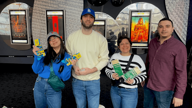 Four people are standing side-by-side in a movie theatre. Two of them are holding up Volkswagen-branded socks. In the background, there movie posters placed along the wall.