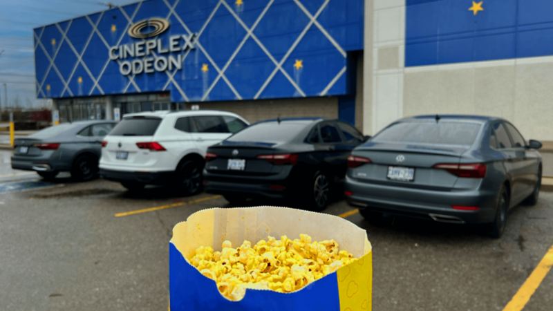 Four Volkswagens are parked next to eachother in the parking lot of a Cineplex movie theatre. In the foreground is a blue bag of movie-theatre popcorn.
